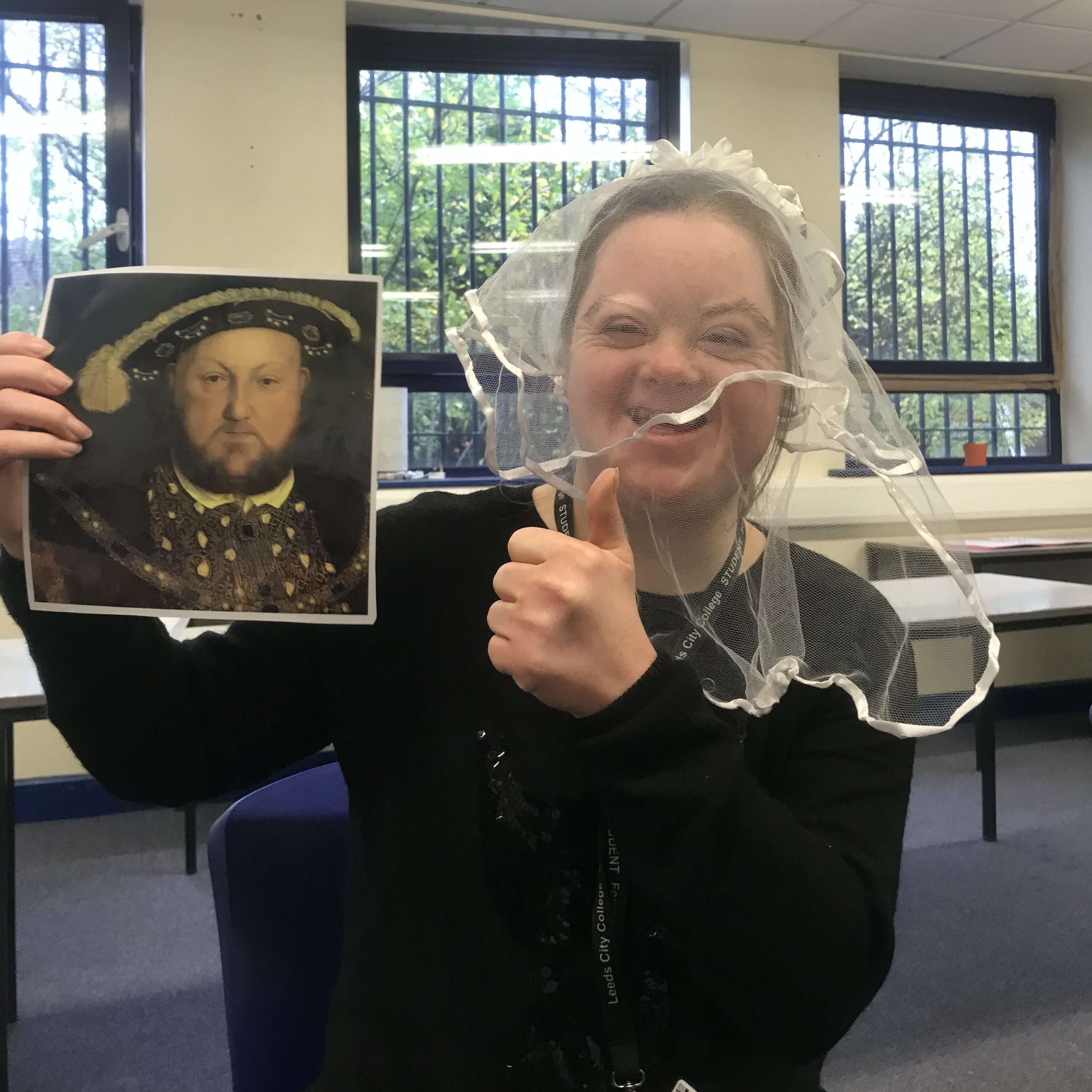 A woman is smiling and doing a thumbs up to the camera. She is wearing a wedding veil and holding up a picture of King Henry VIII.