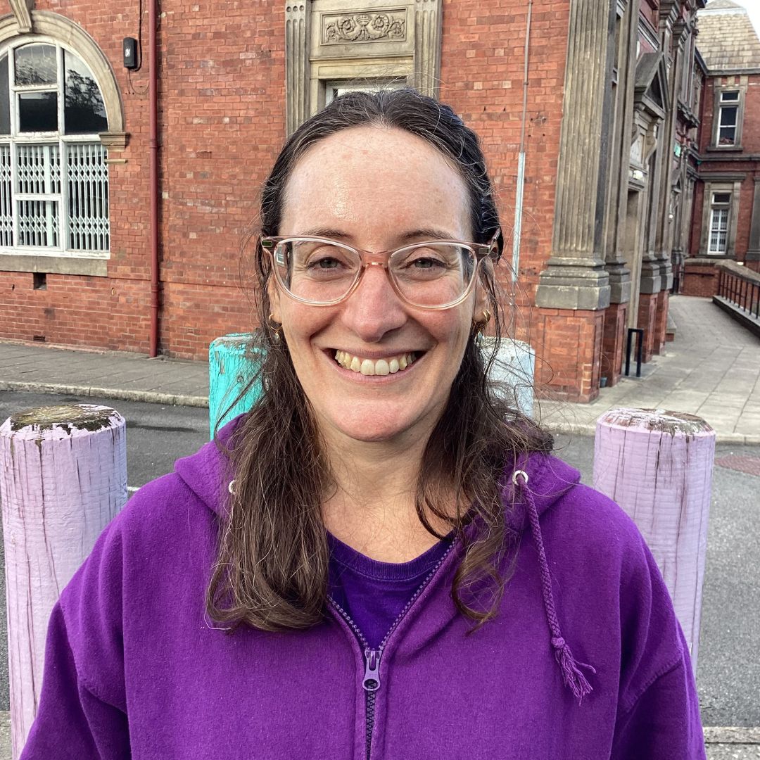 Mati is a woman with medium-length brown hair, glasses and a purple hoody. She is smiling at the camera.